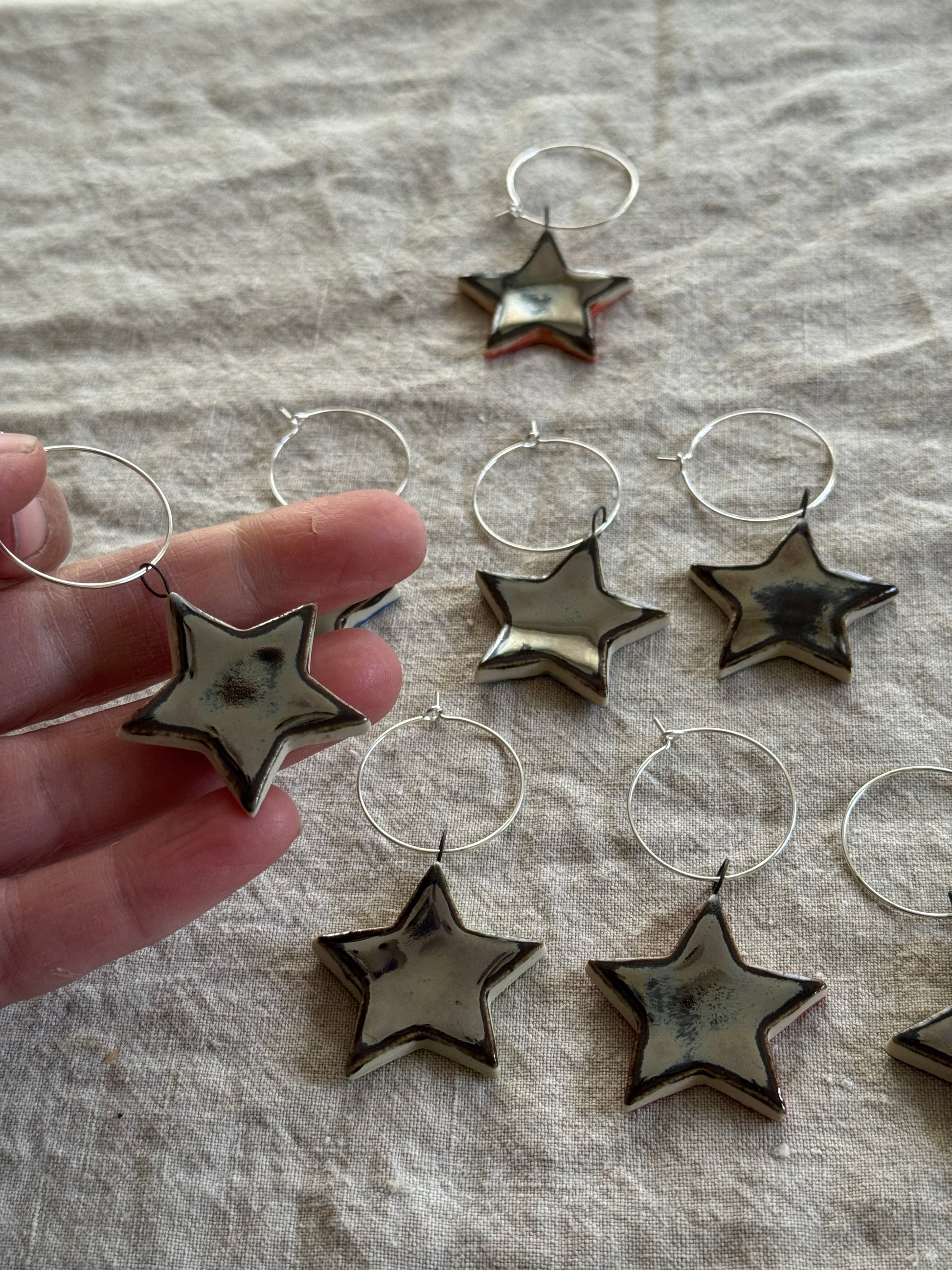 Star-shaped silver ceramic wine charms on silver hoops with a hand holding one, on a textured fabric background.