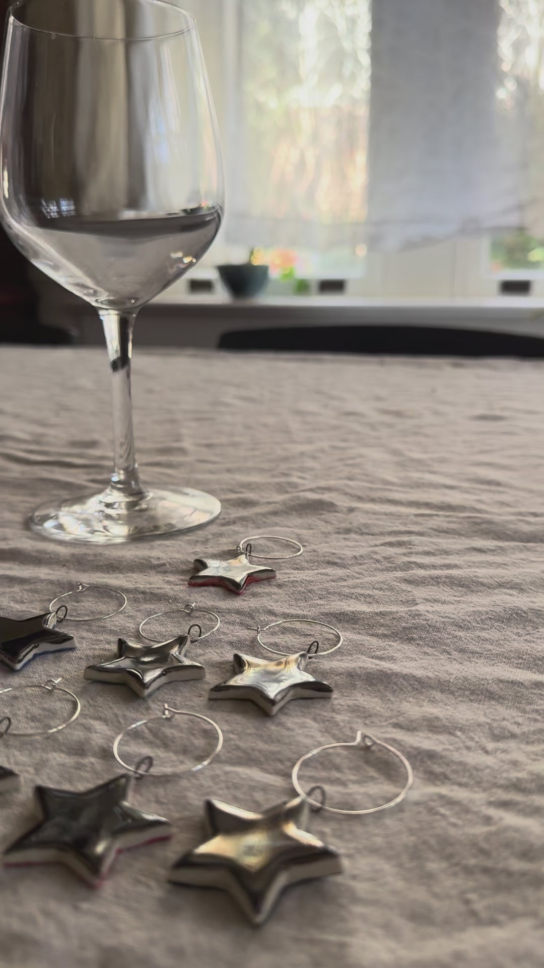 A hand picking up a silver star wine charm and attaching it to an empty wine glass stem, over a textured table cloth. 