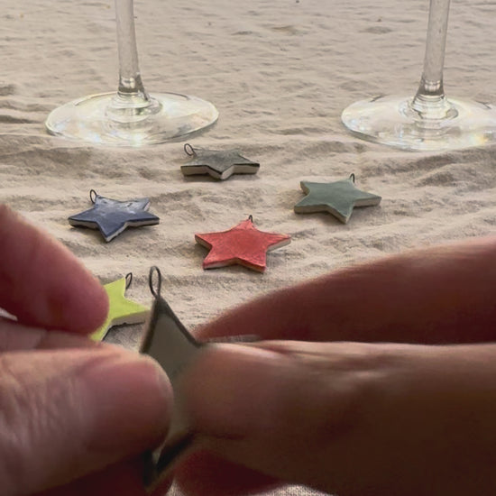 A video of hands connecting a round silver loop to the star shaped drink charms to show how they can be attached to a wine glass stem. more star charms and two wine glasses in the background. 